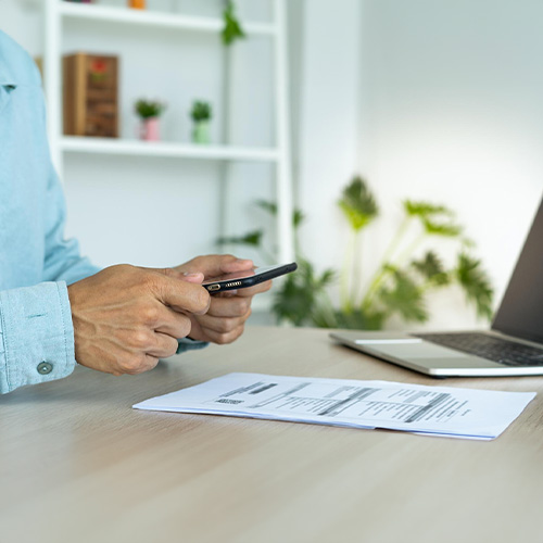A Person Holding a Smartphone in One Hand While Intently Looking at a Laptop Screen, Possibly Managing Online Bill Payment