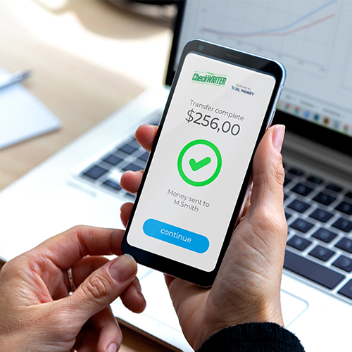 A Person's Hands Holding a Smartphone Display a Payment Methods Successfully Completed Notification to a Recipient, with a Laptop in the Background